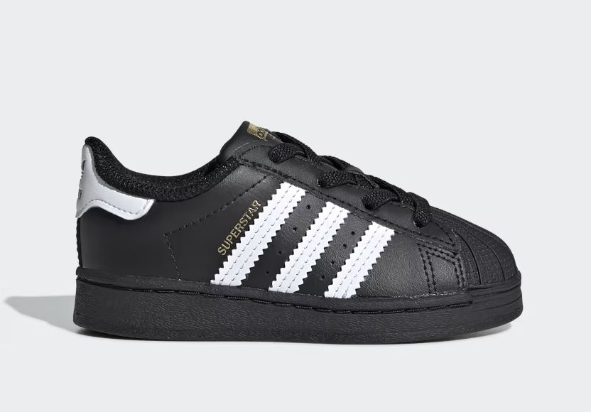 Black adidas toddler shoes, featuring white stripes and gold Superstar text, are shown in profile.