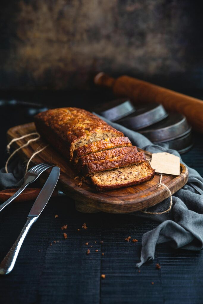Sliced gluten free banana bread on a wooden board, with a knife, fork, and round baking pans in the background.