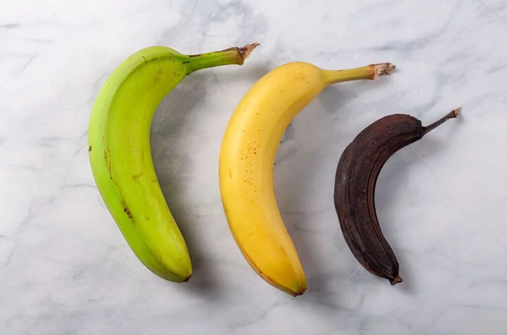 Three bananas: one green, one ripe yellow, and one black banana showing the stages of bananas rotting on a light surface.
