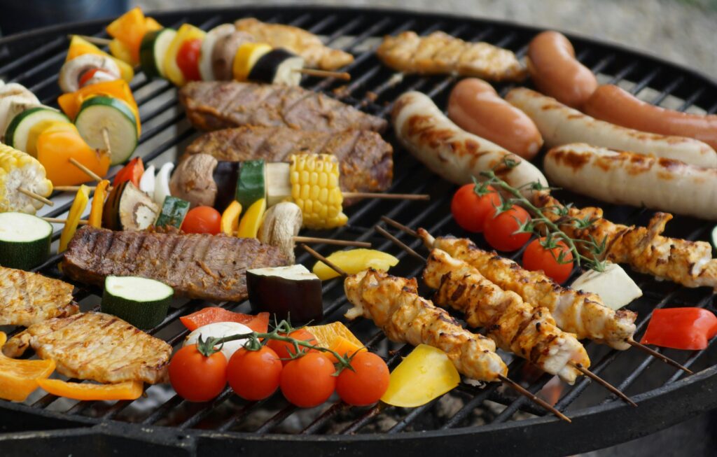 Assorted meats and vegetables, including skewers and sausages, cooking on a charcoal grill for a Gluten Free Life.