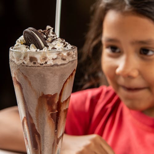 A child excitedly looks at a chocolate milkshake with whipped cream in a glass cup.