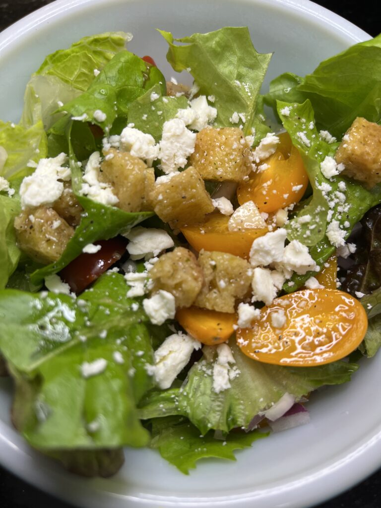 A Panzanella Salad with leafy greens, yellow tomatoes, croutons, and crumbled feta cheese in a white bowl.