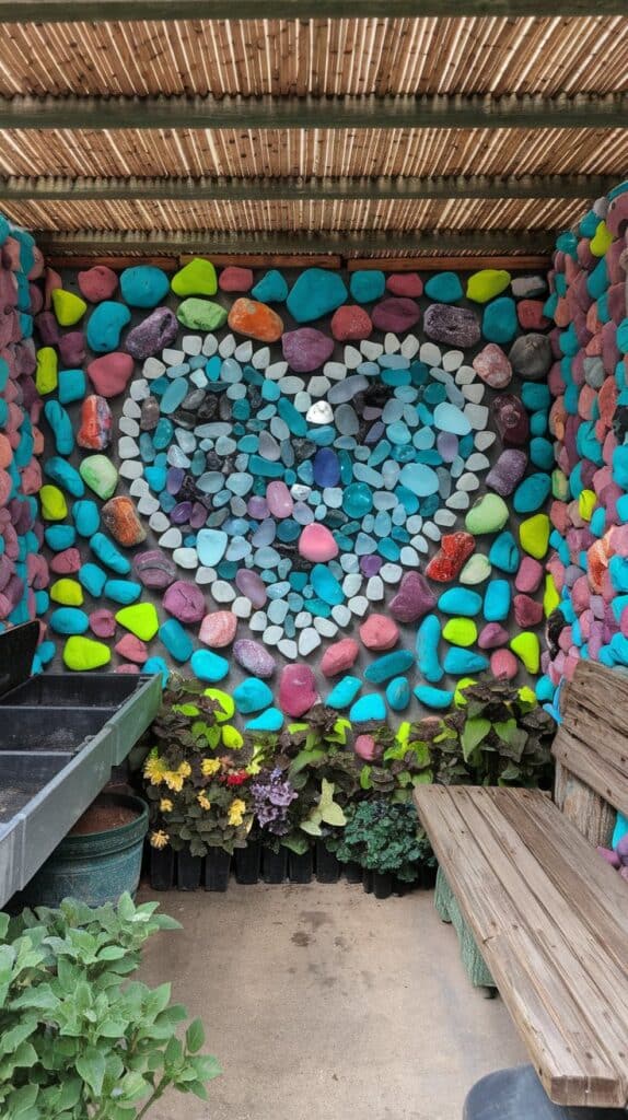 A wall covered in colorful painted rocks, inspired by DIY Sea Glass and Rock Mosaic Ideas, forms a heart above a bench.