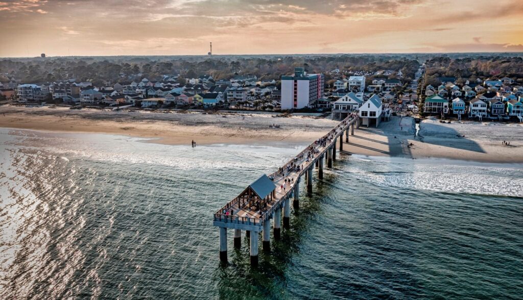 A long pier extends over ocean water toward a sandy beach—one of the Hidden Gems in the South Strand: A Local’s Guide.