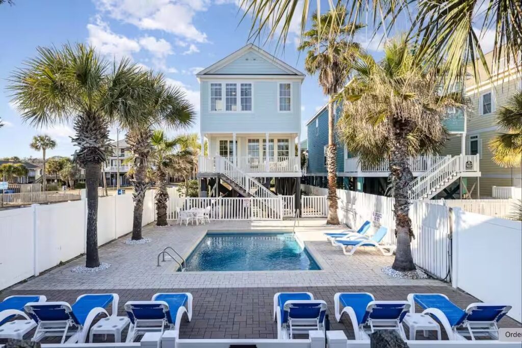 A pool with blue lounge chairs and palm trees awaits at this two-story beach house—your Myrtle Beach vacation guide.