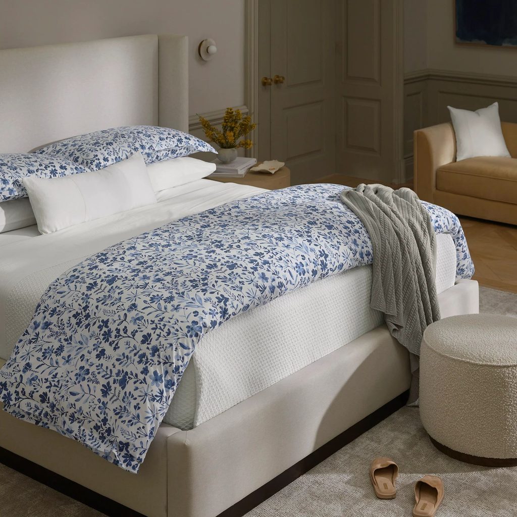 A neatly made bed with blue floral Boll & Branch bedding, a gray throw, beige chair, and slippers on a light carpeted floor.