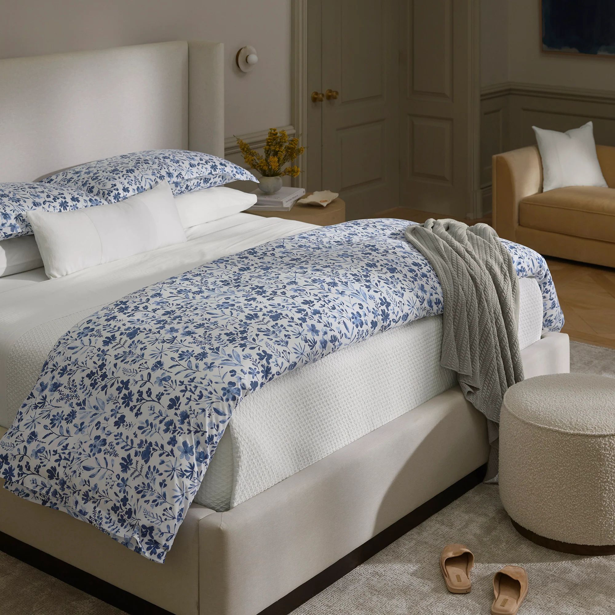 A neatly made bed with blue floral Boll & Branch bedding, a gray throw, beige chair, and slippers on a light carpeted floor.