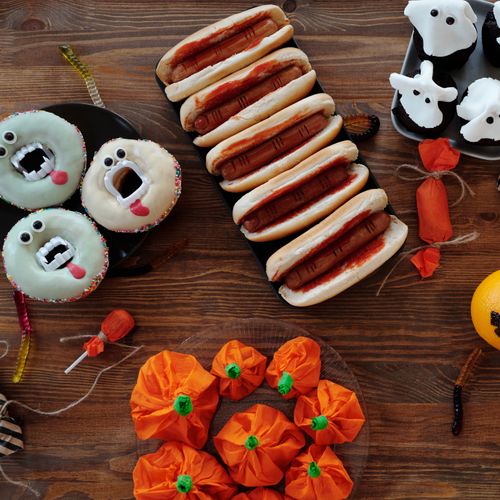 Festive Halloween treats including ghost cupcakes, spooky hot dogs, and monster donuts on a wooden table.