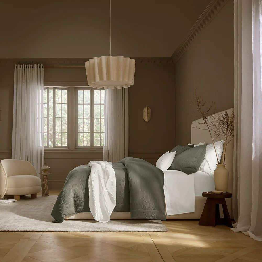 Modern bedroom with green Boll & Branch bedding, a beige chair, large window, sheer curtains, and sculpted pendant light.