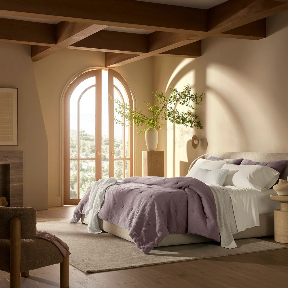 Sunny bedroom with arched window, beige walls, wooden beams, and bed with purple Boll & Branch bedding and potted plant.