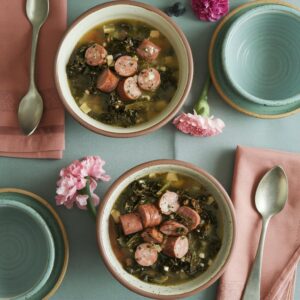 Two bowls of kale and sausage soup on a table set with napkins, spoons, empty bowls, and flowers.